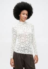 Mujer con peinado afro que lleva una blusa de encaje floral de manga larga blanca y pantalones de cintura alta marrón oscuro, de pie contra un fondo blanco.