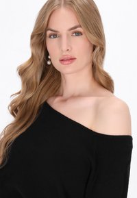 Pulover negru, cu umerii descoperiți și o textură moale. Model cu păr ondulat, de lungime până la umeri și cercei din perle. Fundal neutru.