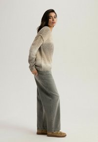 Przesadnie duży beżowy sweter dzianinowy z prążkowanymi mankietami, w połączeniu z luźnymi szarymi spodniami z wysokim stanem i brązowymi butami w stylu mokasynów.