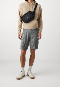 Sudadera beige, pantalones cortos de carga grises con bolsillos laterales, bolsa cruzada negra y zapatillas blancas con suela de goma. Diseño simple y utilitario.