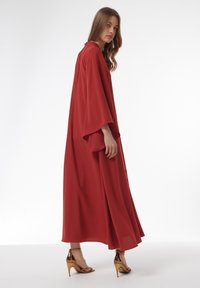 Rode maxi-jurk met brede mouwen, vloeiende silhouet en een gladde textuur. Gecombineerd met glanzende gouden hoge hakken.