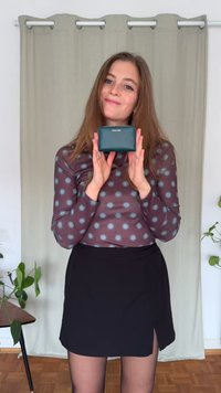 Sieviete ar gariem brūniem matiem tur rokā mazu tirkīzzilu Calvin Klein maks, stāvot telpās priekšā bēša aizkara un augiem.