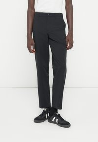 Person iført sorte bukser med lige ben, sorte sneakers med hvide striber og en hvid ærmeløs top mod en ensfarvet baggrund.