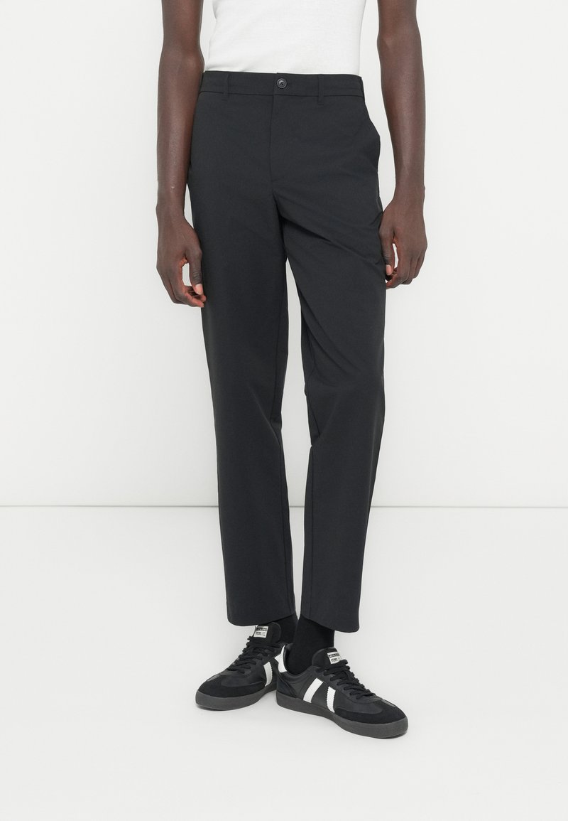 Person iført sorte bukser med lige ben, sorte sneakers med hvide striber og en hvid ærmeløs top mod en ensfarvet baggrund.