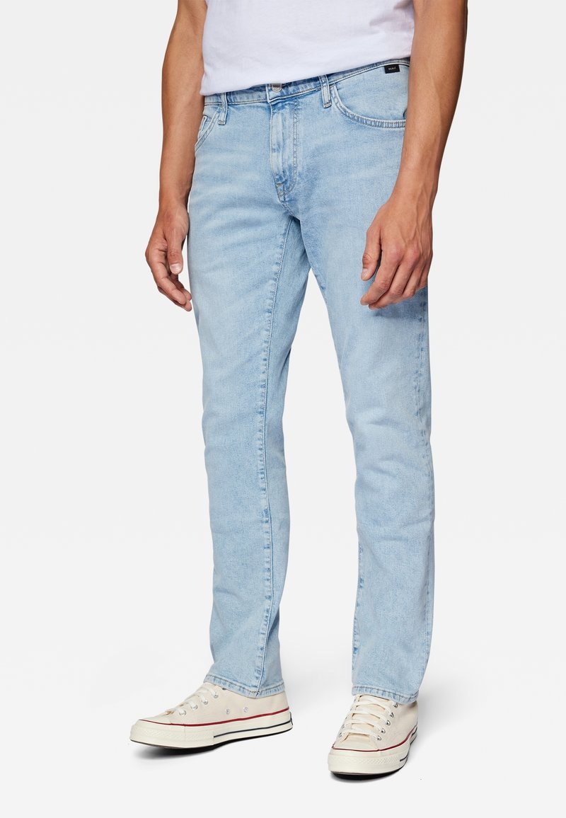 Homme portant un jean bleu clair, des baskets blanches et un t-shirt blanc, debout devant un fond uni et clair.