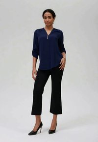 Navy plava bluza na patentni zatvarač s dugim rukavima, uparena s crnim skraćenim hlačama i crnim šiljastim potpeticama. Glatki materijal s ležernim krojem.