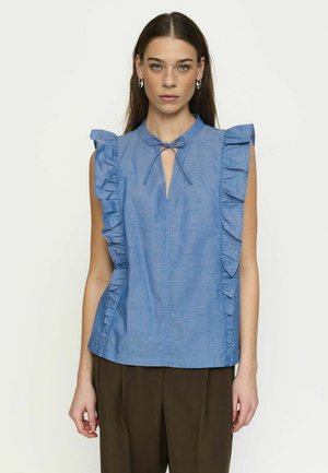 Noor naine pikkade tumejuustega, hõbedaste rõngaskõrvarõngastega, seljas sinine volangidega varrukateta pluus lipsuga kaelusel ja tumedat pruuni värvi püksid.