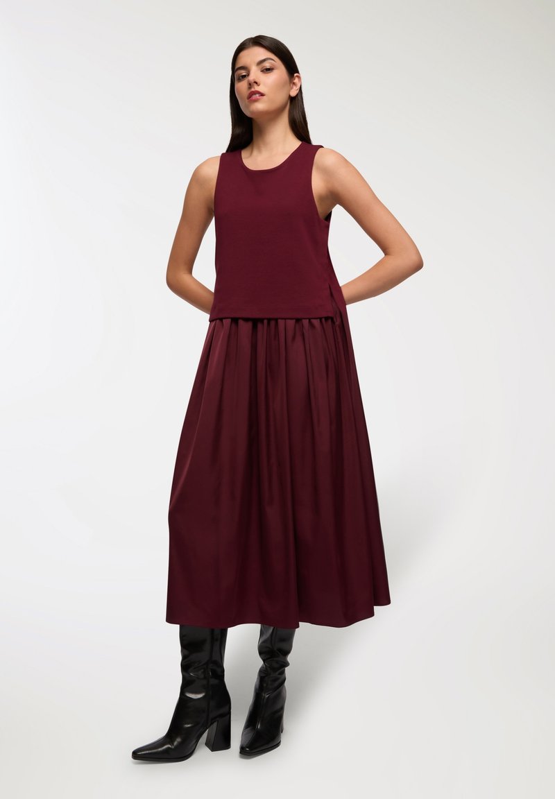 Bordowa top bez rękawów w połączeniu z plisowaną bordową spódnicą o gładkiej teksturze i wysokim stanie, uzupełniona czarnymi botkami na obcasie.