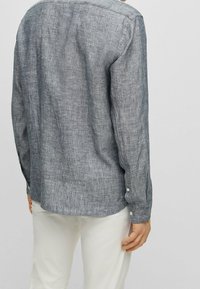 Grå linneskjorta med långa ärmar, knäppta manschetter, rak fåll och en texturerad tyg. Bakifrån syns en enkel krage och rena linjer.