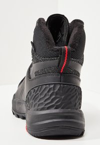 Mustapohjaiset korkeavartiset saappaat, joissa on teksturoitu kumipohja, verkkopinta ja pehmeä, eristetty varsi, jossa on punainen yksityiskohta.