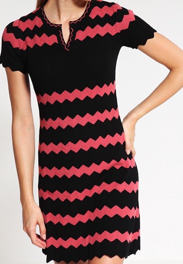Femme portant une robe noire à manches courtes avec des rayures zigzag roses, des bords festonnés aux manches, à l’ourlet et à l’encolure, debout, la main sur la hanche.