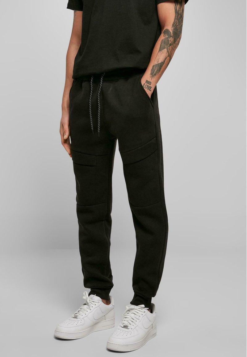 Sorte joggebukser lavet af blødt materiale, med sidelommer og en tapered fit, parret med hvide sneakers.