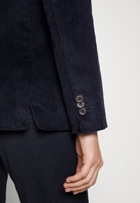 Námořnický kordový blazer s přiléhavým střihem. Obsahuje tři knoflíky na manžetách a texturované vertikální žebrování po celé látce.