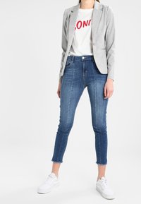 Femme portant un blazer gris, un t-shirt blanc avec un texte rouge, un jean skinny bleu coupé au-dessus de la cheville et des baskets blanches, debout sur un fond blanc.