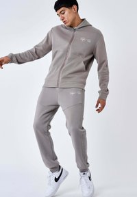 Sweat à capuche gris zippé avec logo, associé à un pantalon de jogging gris assorti avec des accents latéraux et une texture lisse. Porté avec des baskets blanches.