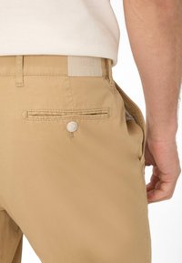 Beige broek met een achterzak, voorzien van een knoopsluiting en een tekstiel label. De stof is glad met een lichte glans.