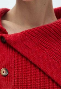 Cardigan en tricot rouge avec une finition texturée, doté d'un large col et de deux gros boutons foncés à l'avant pour la fermeture.