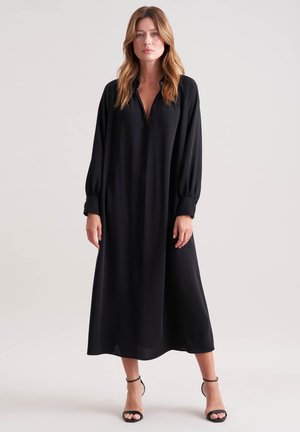 Femme se tenant debout, portant une robe midi noire à manches longues et décolleté en V, avec des escarpins à strap cheville noirs, sur un fond uni.