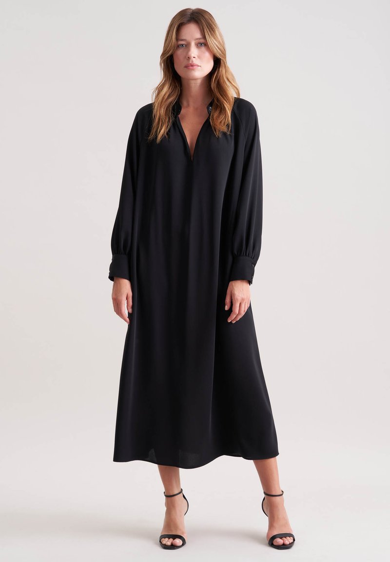 Donna in piedi che indossa un lungo vestito midi nero a maniche lunghe e scollatura a V, abbinato a tacchi neri con cinturino alla caviglia, su uno sfondo neutro.