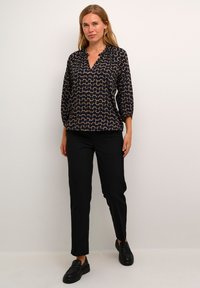 Marineblaue Bluse mit abstraktem beigen Muster, V-Ausschnitt, Dreiviertelärmel, kombiniert mit schwarzen Hosen und schwarzen Slippern.