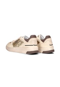 Beige sneakers hebben een glitterpaneel, bruine accenten en een gestructureerde zool. Het ontwerp bevat decoratieve stiksels en een vetersluiting aan de voorkant.