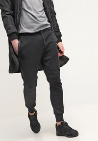 Black jogger pants with elastic waistband and drawstring, featuring side pockets and tapered ankle cuffs, paired with black athletic shoes.