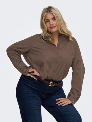 Blond kvinde iført en brun bluse med lange ærmer med snørebånd på manchetterne og mørkeblå jeans med et brunt bælte, der poserer med hånden på hoften.