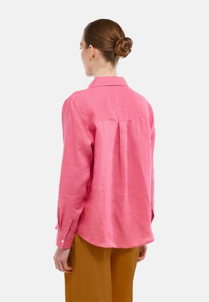 Femme aux cheveux bruns attachés en chignon, portant une chemise rose à manches longues et un pantalon couleur moutarde, vue de dos sur un fond blanc.