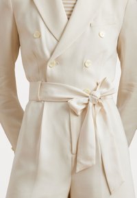 Κρέμα διπλή σακάκι με κίτρινους κουμπιά, προσαρμοσμένη εφαρμογή και μεσοφόρι που διαθέτει ζώνη με κόμπο. Λείος ύφασμα και διακριτική γυαλάδα.