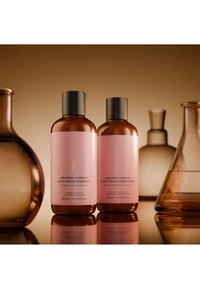 Zwei braune Flaschen mit pinkfarbenen Etiketten, beschriftet mit Monpure Ceramide Complex Bond Repair Shampoo und Conditioner, umgeben von bernsteinfarbenen Laborflaschen aus Glas.