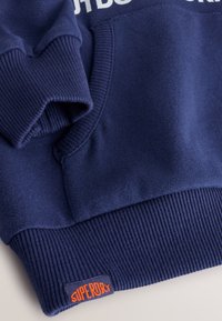 Navy blue sweatshirt with ribbed cuffs and hem. Features a textured logo label in orange, alongside a white printed text design on the front.