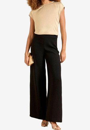 Kvinde iført beige top med korte ærmer, højtaljede vide sorte bukser, gyldent kuvertetui og gyldne åbne hæle mod en hvid baggrund.