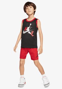 Czarny siateczkowy tank top z czerwonym obszyciem i graficznym logo, zestawiony z czerwonymi szortami z czarnym logo, noszony z białymi trampkami.