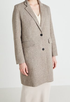 Femme portant un manteau en laine taupe mi-genoux avec deux boutons noirs et des poches à rabat sur une robe de couleur claire.