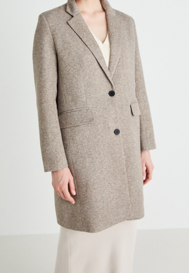 Femme portant un manteau en laine taupe mi-genoux avec deux boutons noirs et des poches à rabat sur une robe de couleur claire.
