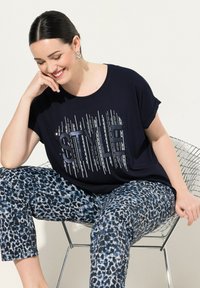Námořnické tričko s krátkým rukávem a metalickým nápisem "STYLE", zkombinované s croptop kalhotami v modro-šedém leopardím vzoru, sedící na drátové židli.