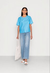 Top azul de manga curta, com um corte descontraído e textura suave, combinado com jeans de perna larga azul-claro. Sandálias brancas de slide e uma mala de tiras.