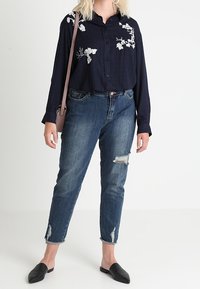 Námořnická košile s knoflíky a bílou květinovou výšivkou, kombinovaná s otrhanými džínami. Modelka má na sobě černé slip-on boty a nese béžovou tašku.