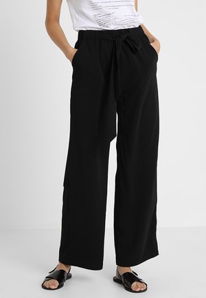 Person wearing black wide-leg pants with a tied fabric belt, white printed t-shirt, and black open-toe slip-on sandals, hands in pockets.