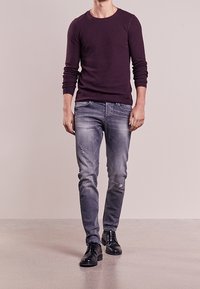 Mörklila långärmad skjorta, grå slitna jeans och svarta snörskor. Modellen står mot en ljus bakgrund, med synlig betonggolvtextur.