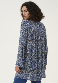 Tunique florale en crème, bleu et noir. Présente des manches longues, un dos plissé et une coupe fluide avec des motifs complexes partout.