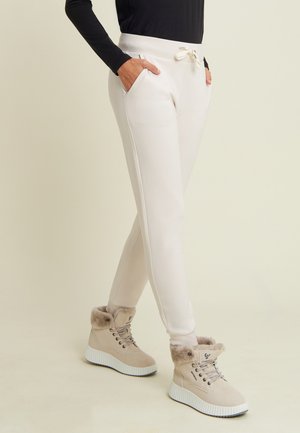 Persona che indossa pantaloni jogger bianchi con coulisse, stivaletti beige foderati in pelliccia, e una maglia nera a maniche lunghe, in piedi con le mani nelle tasche.