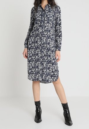 Femme portant une robe fleurie bleu marine, longue jusqu’aux genoux, à manches longues, associée à des bottines noires brillantes, debout devant un fond uni.