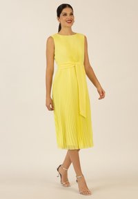 Faltenrockkleid in Gelb mit Midi-Länge, Rundhalsausschnitt und einem gebundenen Taillendetail. Silberne Riemchen-Absatzsandalen vervollständigen das Outfit.
