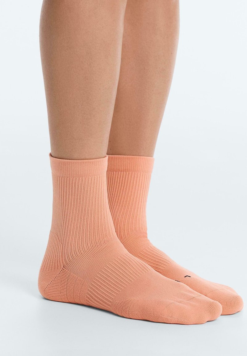 Beine mit gerippten, knöchellangen, pfirsichfarbenen Socken, die auf einer glatten, hellen Oberfläche vor einem schlichten Hintergrund stehen.