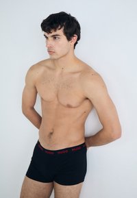 Boxers en coton noirs avec une ceinture rouge "HUGO", portés par une silhouette masculine aux cheveux courts et foncés, se tenant devant un fond clair.
