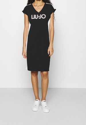 Kvinde iført en sort knælængde kjole med hvide sidepaneler og logoet "LIU JO", kombineret med hvide sneakers og en guldkæde.