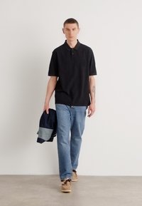 Sort polo-shirt med diskret branding, parret med lyseblå jeans. Figuren holder en mørk jakke og har beige sko på. Neutral baggrund.