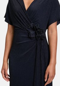 Marineblaues Kleid mit gerippter Textur, V-Ausschnitt und kurzen, schwingenden Ärmeln. Es hat eine geraffte Blumenakzent an der Taille und einen fließenden Fall.