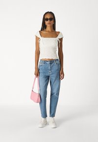 Biely textúrovaný crop top s krátkymi rukávmi, modré džínsy s vysokým pásom, ružová kabelka a biele platformové tenisky. Slnečné okuliare sú súčasťou.